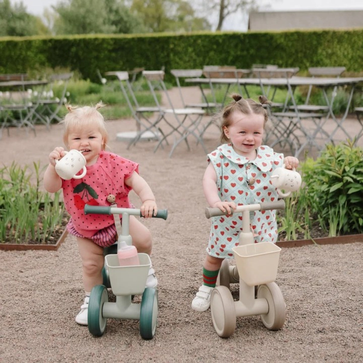 two toddlers riding Mushie Mini Toddler Bikes outdoors in a garden