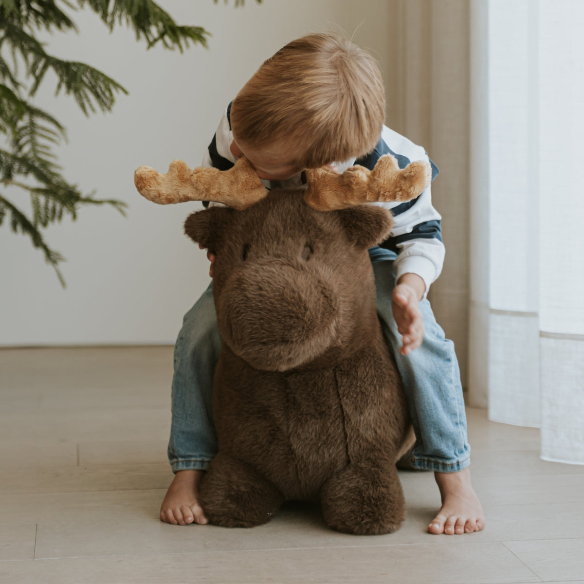 a young child sitting on a Wild & Soft Bouncy Moose plush animal toy for children aged 3+ next to christmas tree