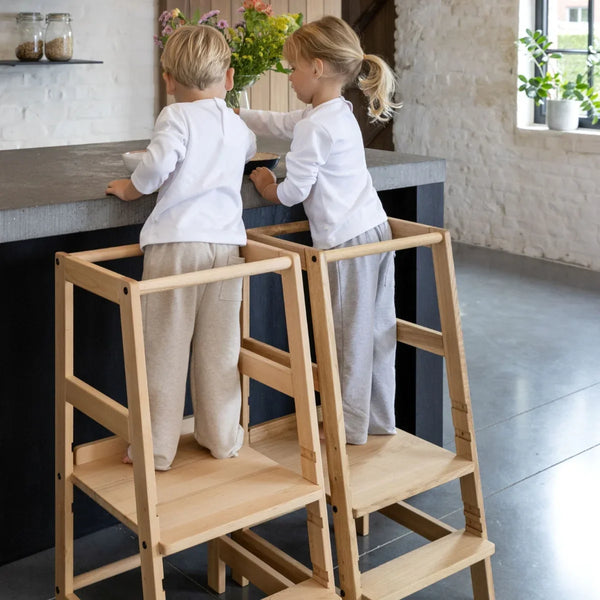 two young children standing on wooden learning towers in modern kitchen