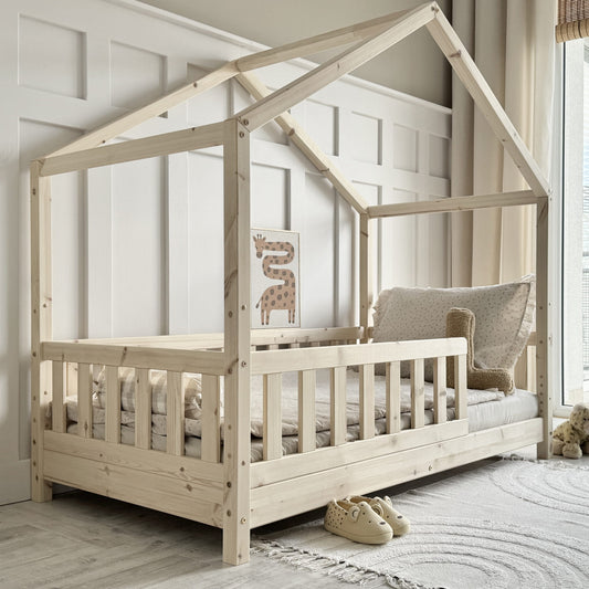 Wooden children's house shaped bed with bed guard in a room with a window and rug.