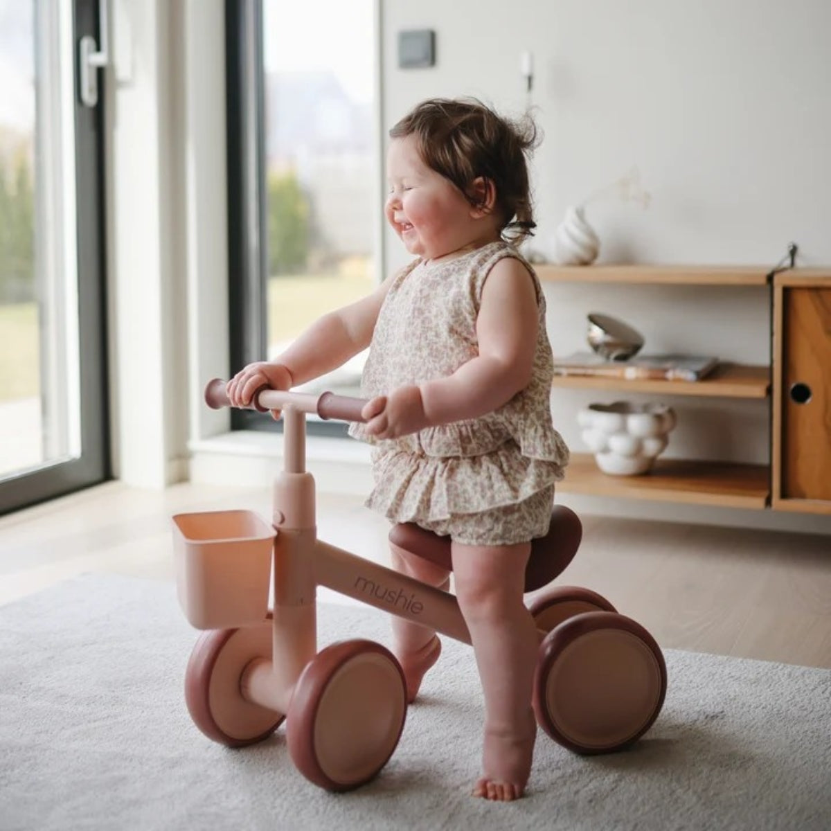Child riding a pink mushie balance bike indoors with a neutral background