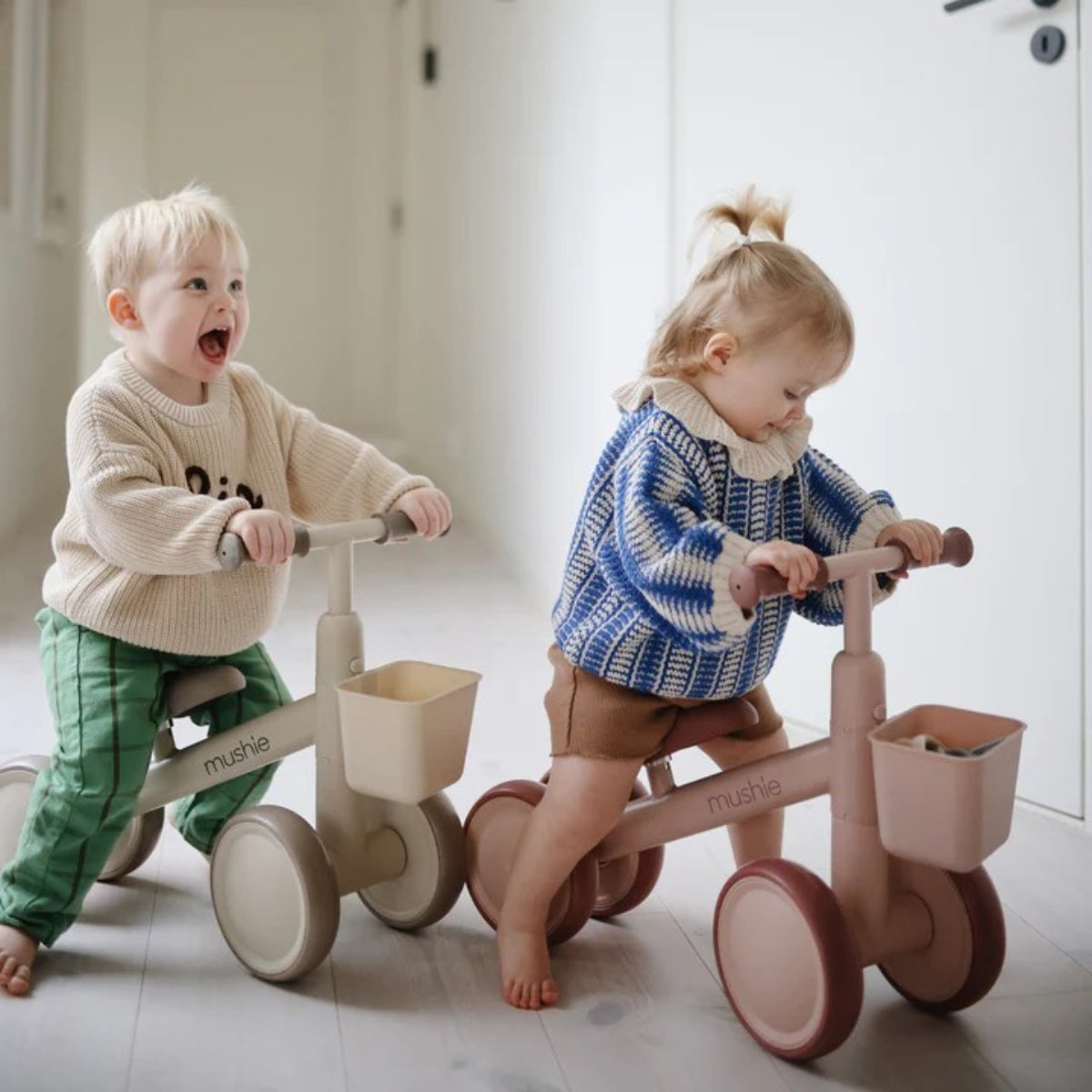 Two children playing with mushie mini toddler balance bikes indoors.