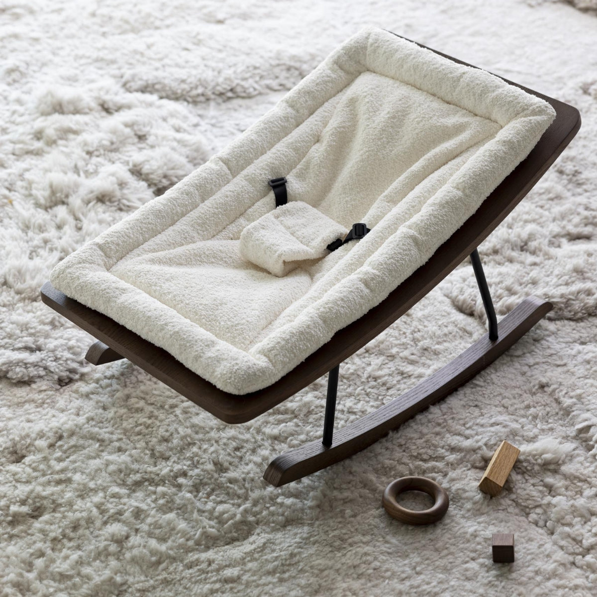 Quax Wooden Rocking Baby Bouncer in Cream bouclé with natural ash dark wooden rockers and 3-point harness on a soft textured rug
