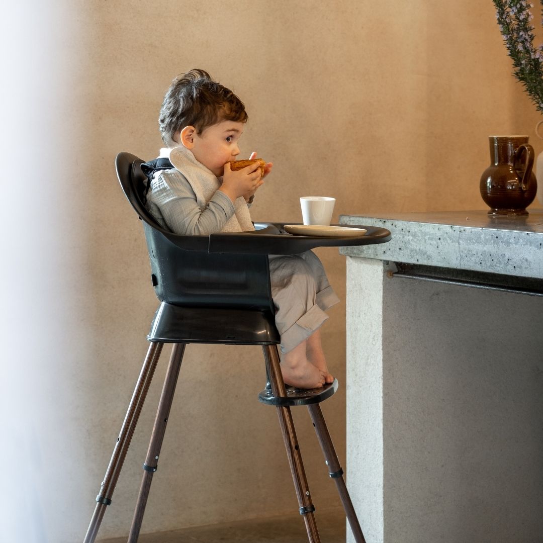 Child sitting in a quax ultimo 3 luxe black walnute high chair eating at a kitchen counter.