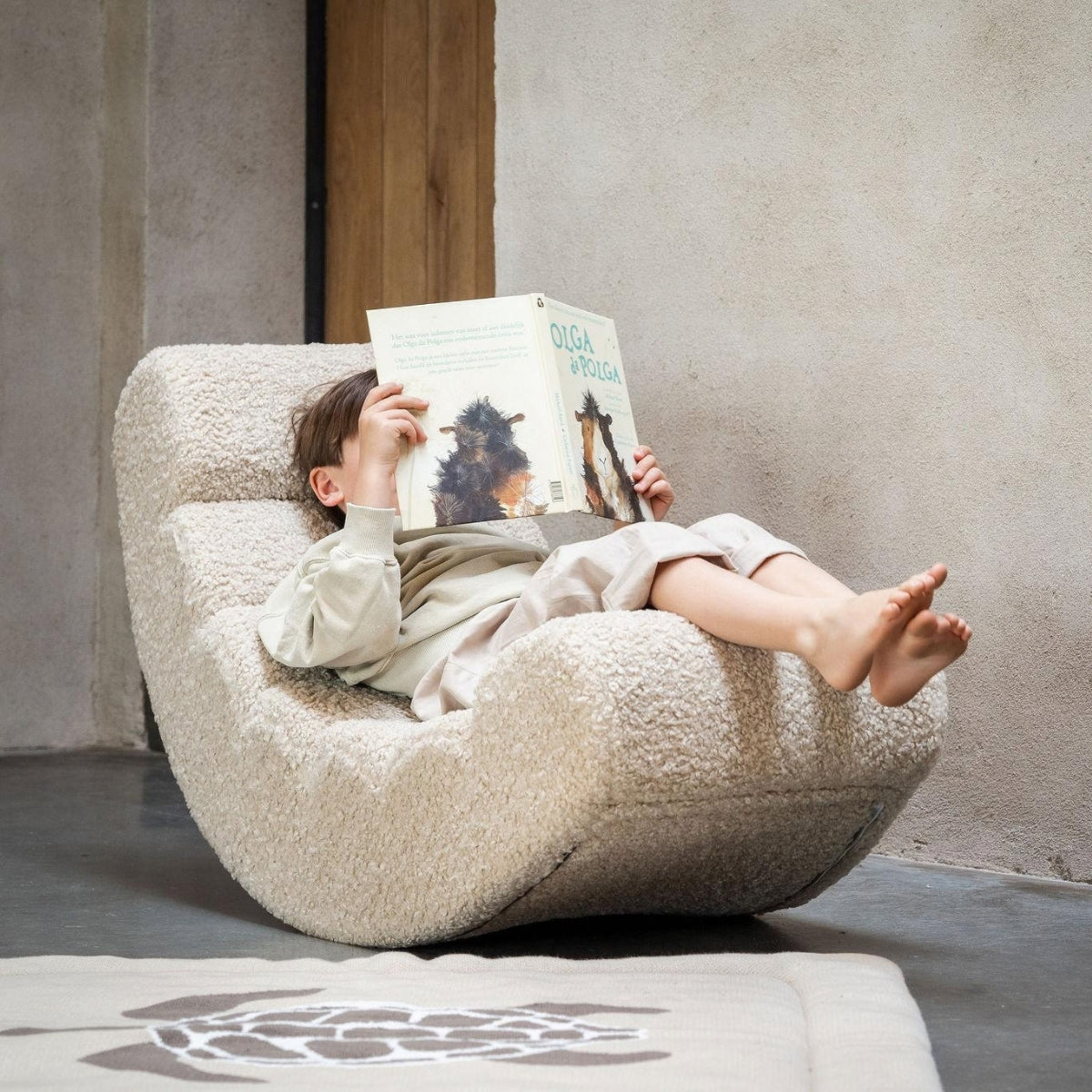 A child lies on a Quax Rocking Banana Chair while reading a book set in a minimalist room 