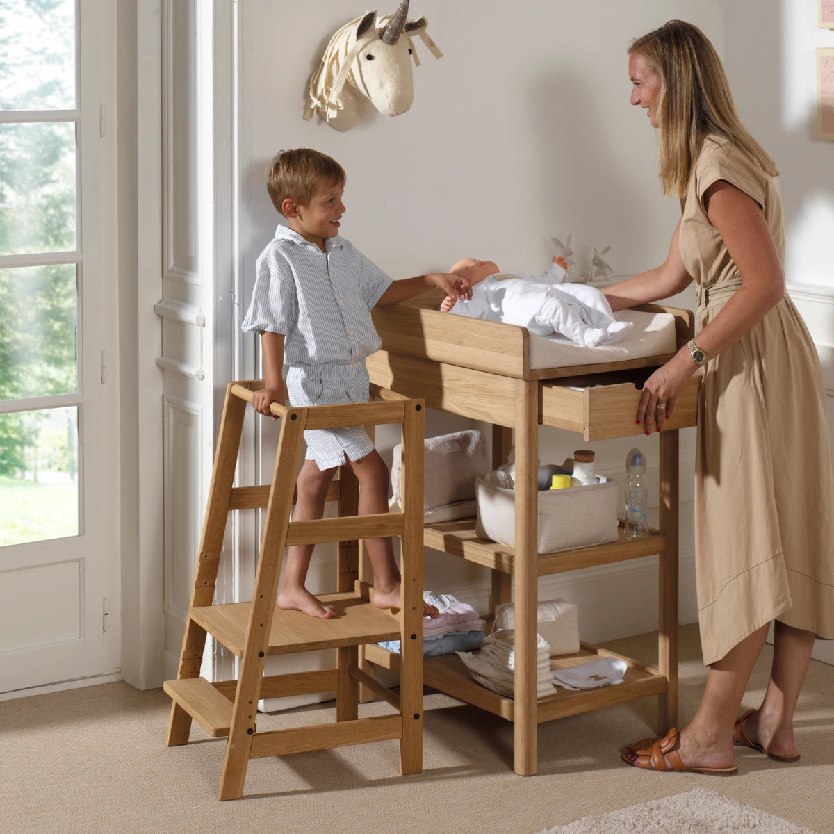 child standing on a solid oak wood Vipack Jaxx Forrest Toddler Learning Tower while his mother stands next to him changing his baby siblings nappy