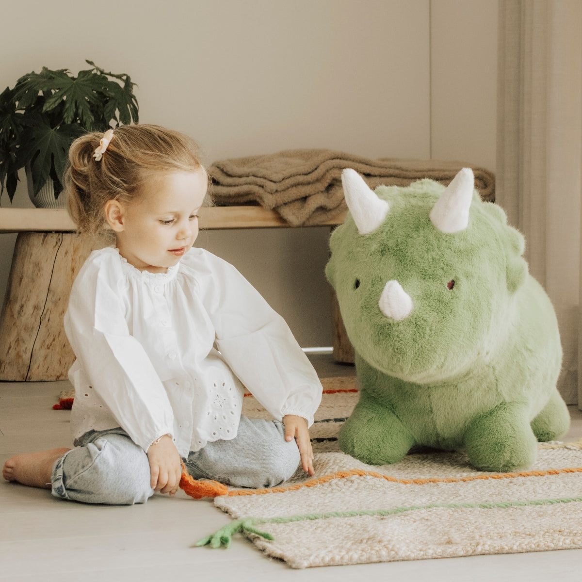 ypung girl sat next to Wild & Soft Bouncy Dino soft plush ride-on dinosaur toy in playroom