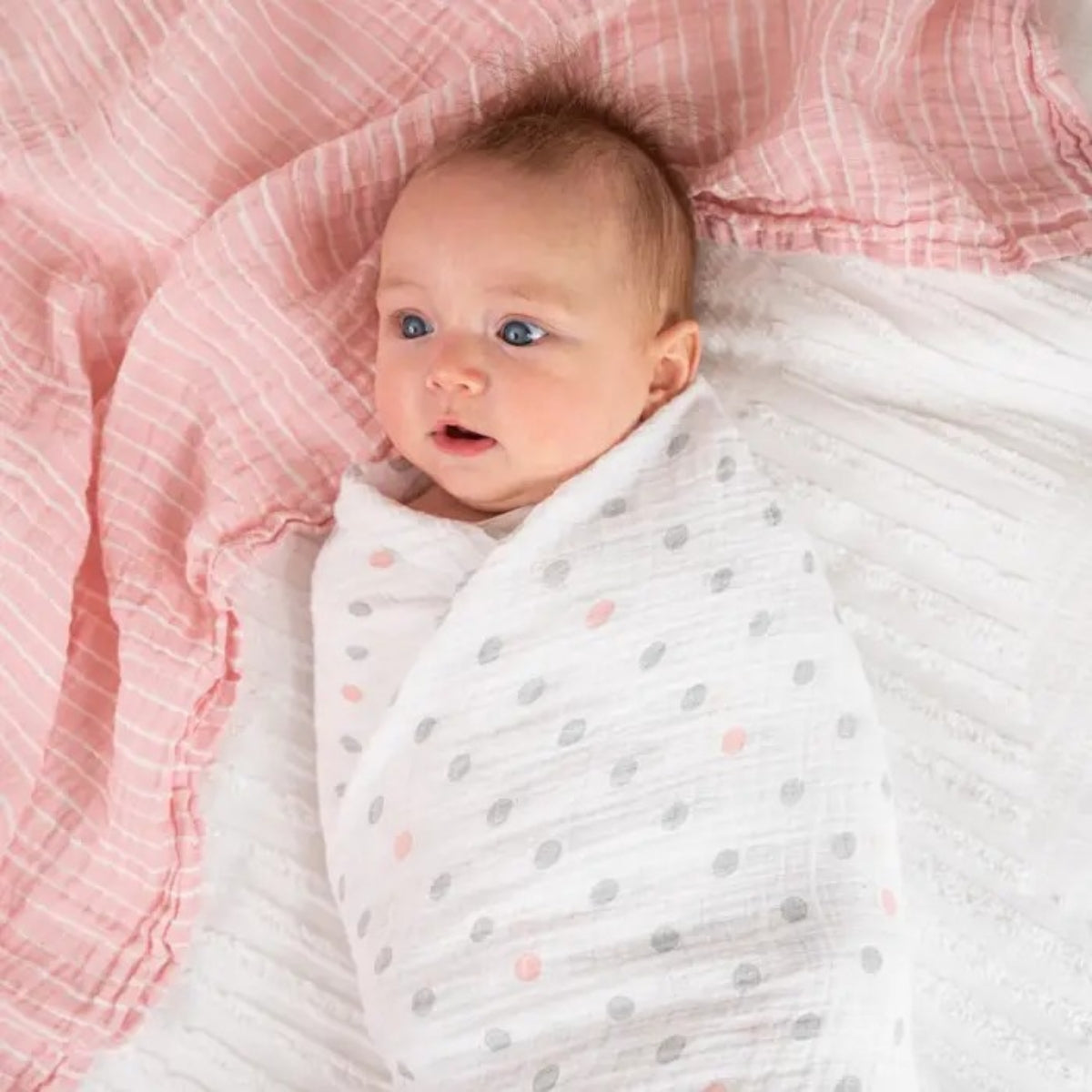Baby wrapped in a white aden + anais cotton muslin blanket with grey and pink polka dots, lying on pink textured fabric.