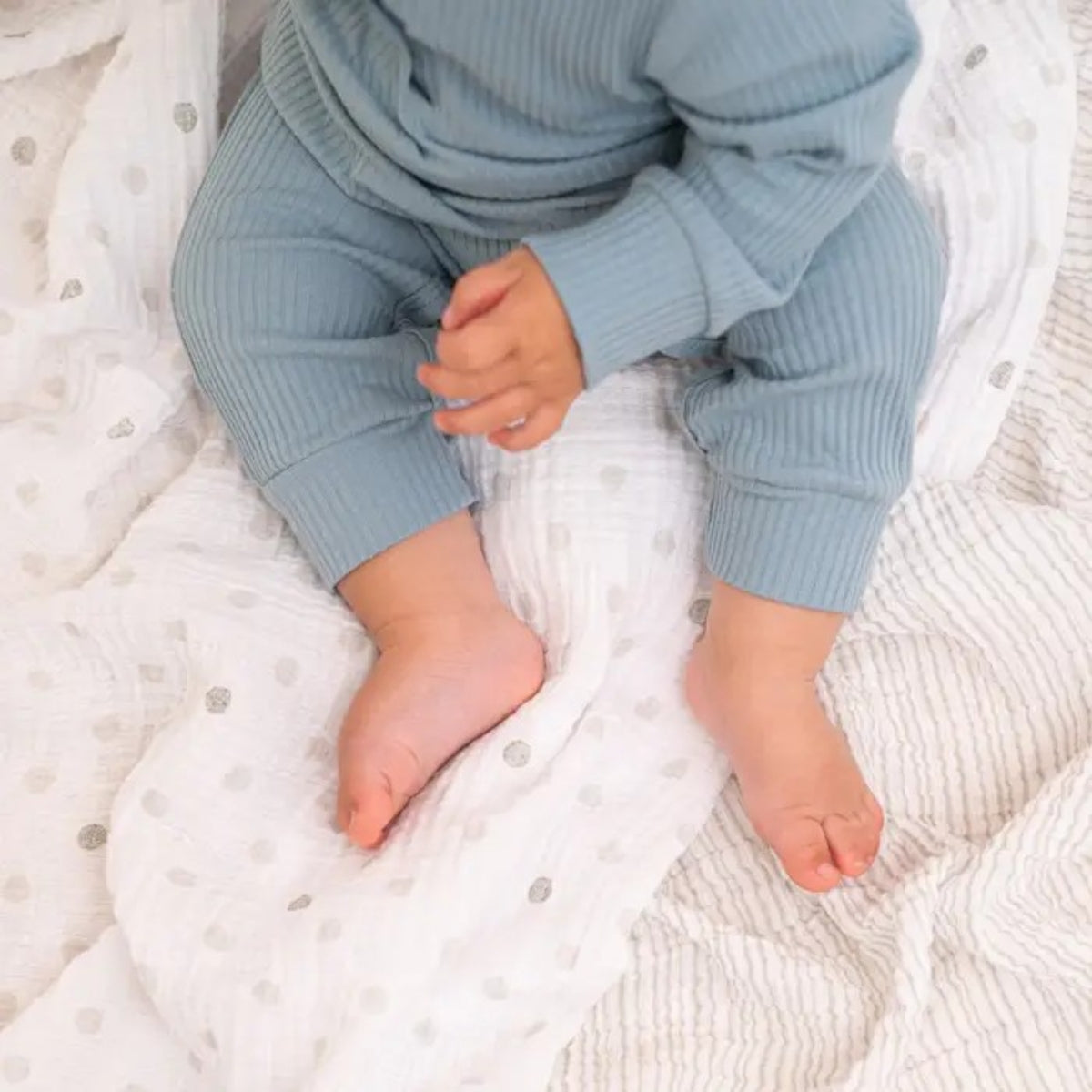 Baby wearing blue ribbed pants sitting on a textured white blanket
