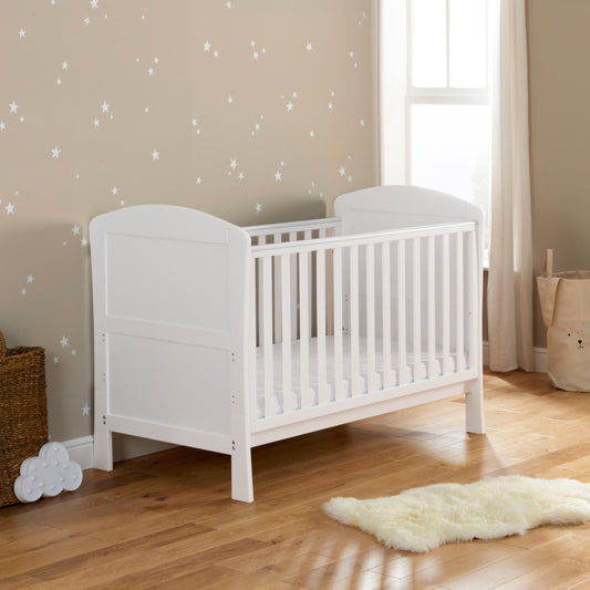 A white Babymore Aston Drop Side Cot Bed with a classic slatted design, situated in a nursery room with a window