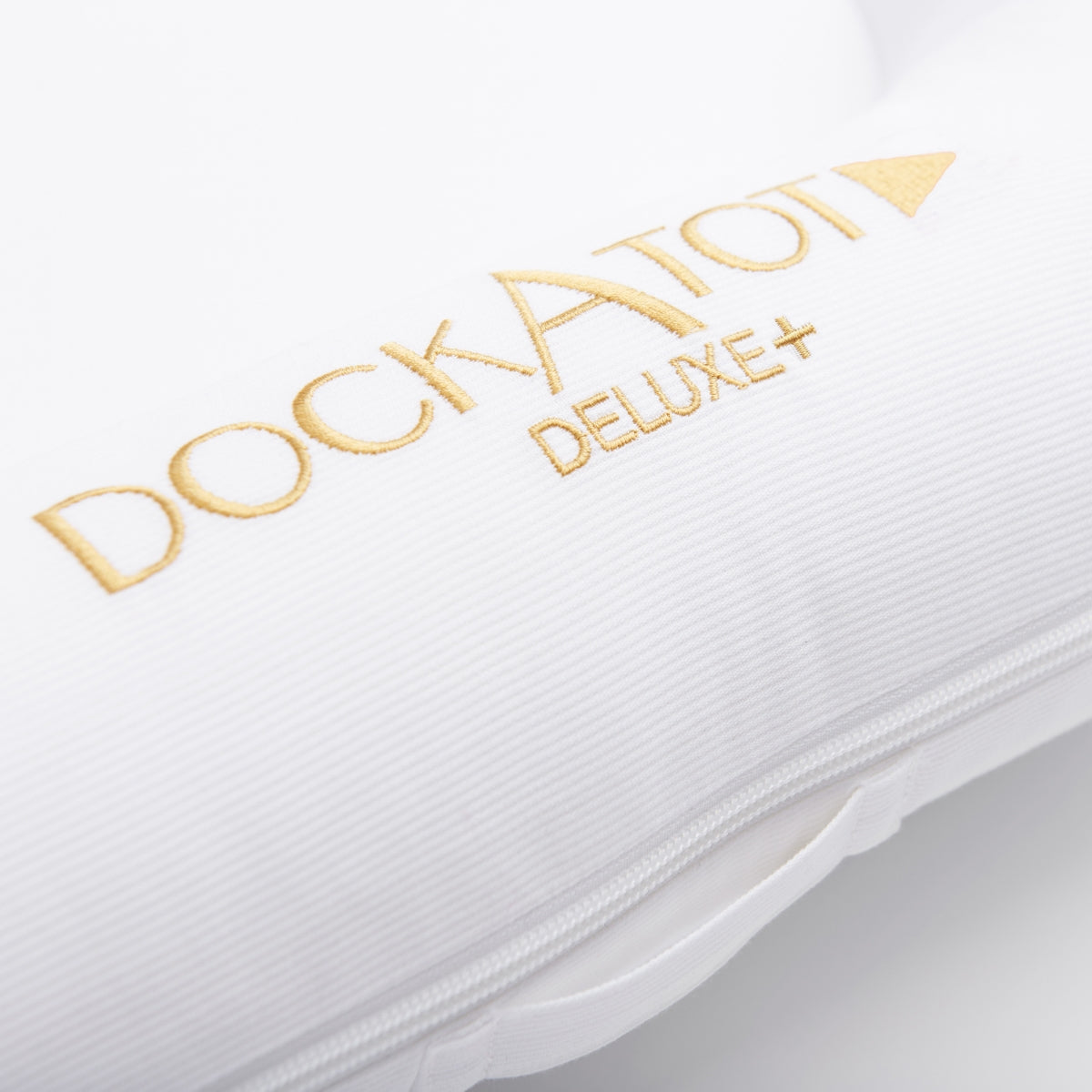 Close-up of a white Dockatot Deluxe+ baby nest with gold embroidered branding and a zipper.
