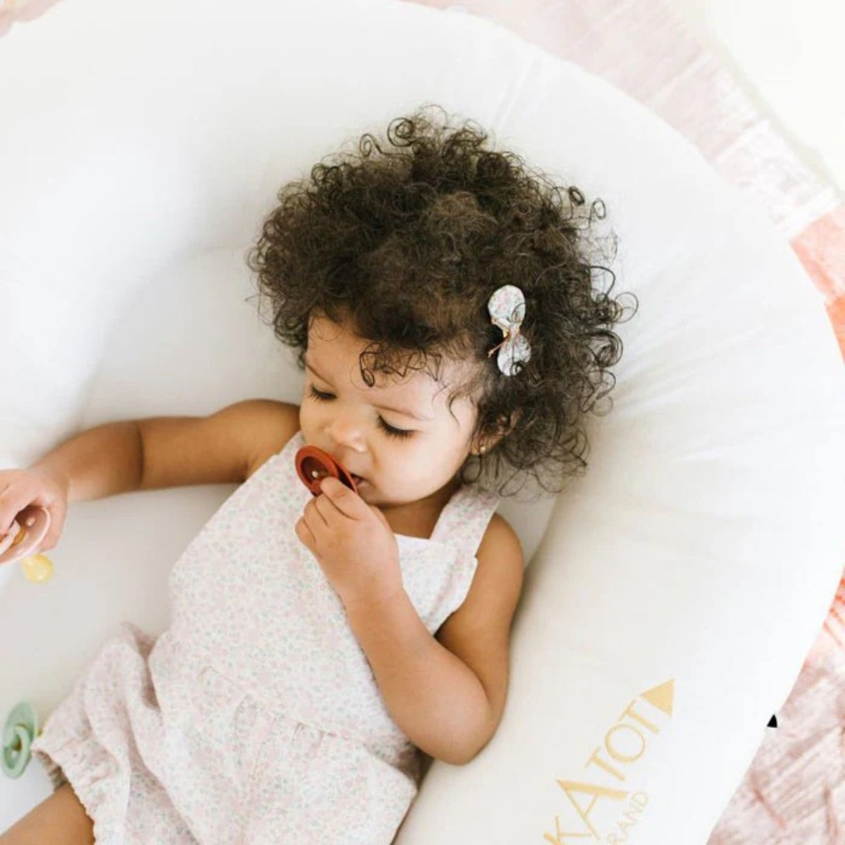 A baby with curly dark hair wearing a light pink floral dress lies on a pristine white DockaTot baby lounger