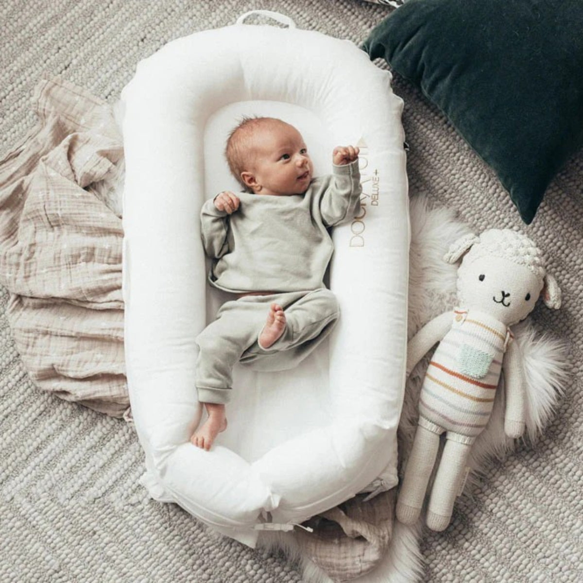baby in a DockATot Deluxe+ Baby Dock Pristine White baby nest with muslin blanket and toy on the floor