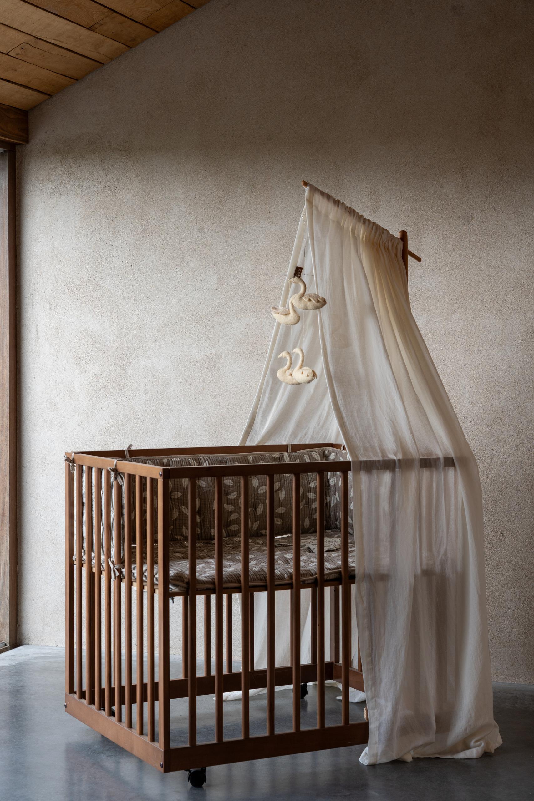 quax butter canopy veil with swan decorations hanging from a wooden frame over a wooden scandi baby playepen against a neutral wall.
