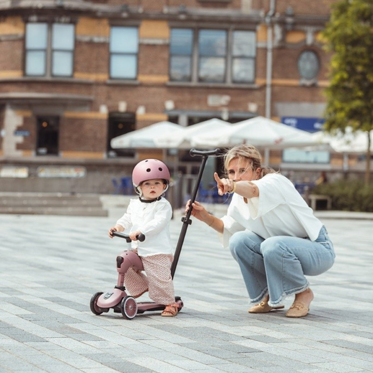 Child on a scoot and ride pink scooter with an adult nearby in an outdoor setting