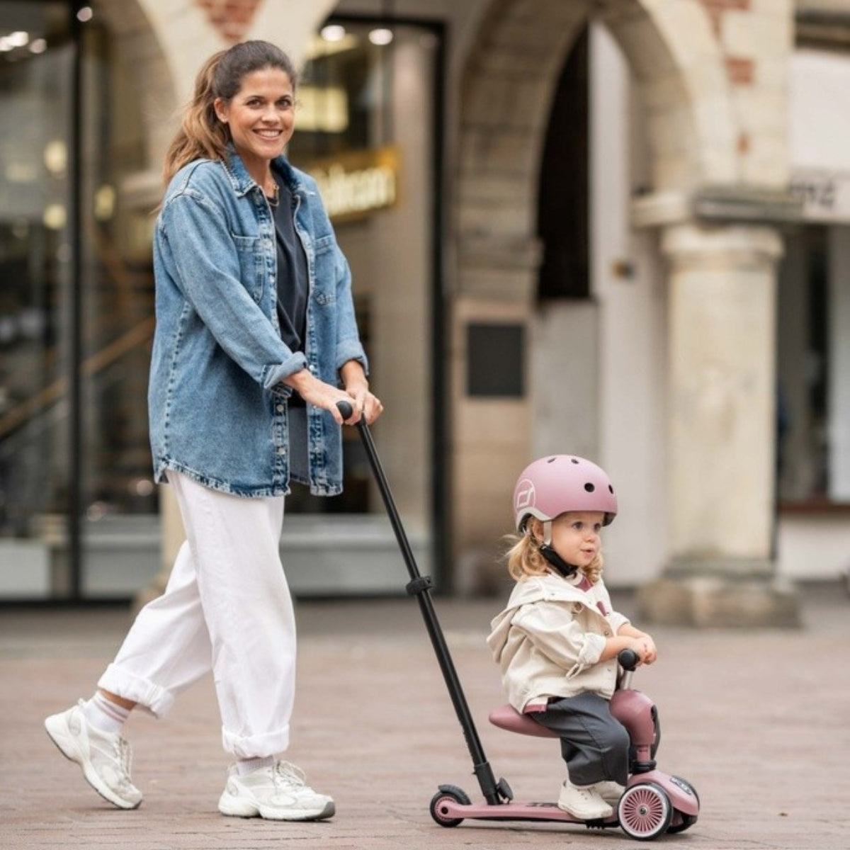 Woman pushing a child on a scoot and ride wildberry scooter in an outdoor setting